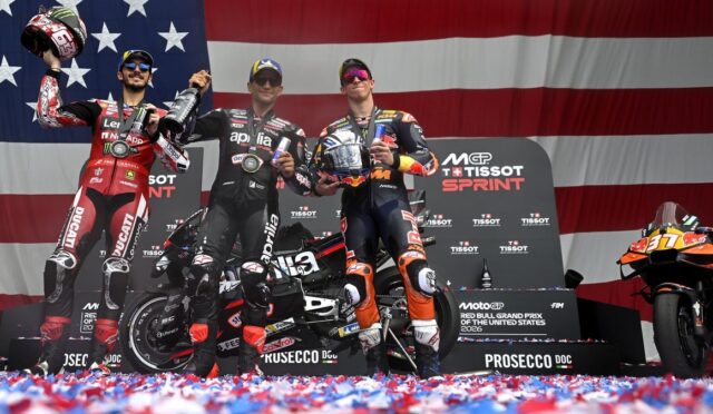 Three MotoGP riders on the winners' podium celebrating with drink bottles, backed by a large American flag banner and sponsor boards. The center rider holds a trophy or helmet, with two others raising their bottles in celebration.