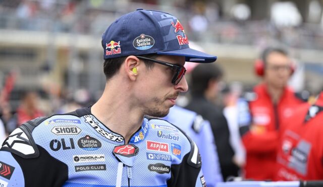 Motorcycle racer in blue suit and cap with sponsor logos, wearing sunglasses and yellow ear protection, in the paddock/garage area.]]