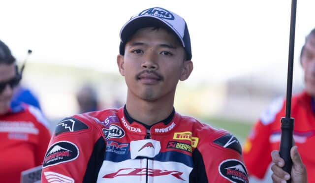 Portrait of a young motorcycle racer in a red Honda racing suit, wearing a blue and white cap, looking at the camera.