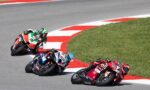 Three motorcycle racers lean into a sharp turn on a racetrack, blue, green, and red bikes close together on the curve.