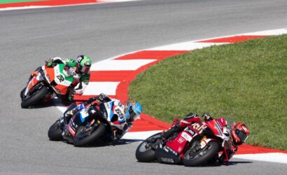 Three motorcycle racers lean into a sharp turn on a racetrack, blue, green, and red bikes close together on the curve.