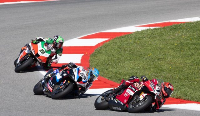 Three motorcycle racers lean into a sharp turn on a racetrack, blue, green, and red bikes close together on the curve.