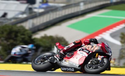 Red racing motorcycle leaning hard into a right-hand bend on a sunny track, rider in matching red suit with sponsor logos visible on the bike’s fairing.