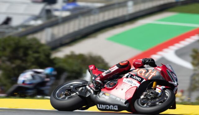 Red racing motorcycle leaning hard into a right-hand bend on a sunny track, rider in matching red suit with sponsor logos visible on the bike’s fairing.