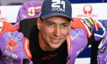 Smiling motorcycle racer in a purple racing suit and cap, leaning toward the camera with sponsor logos visible.
