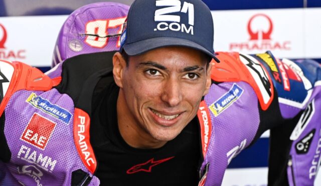 Smiling motorcycle racer in a purple racing suit and cap, leaning toward the camera with sponsor logos visible.
