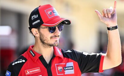 Motorsport team member in a red-black cap and polo with sponsor logos raises his right index finger while wearing sunglasses