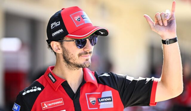 Motorsport team member in a red-black cap and polo with sponsor logos raises his right index finger while wearing sunglasses