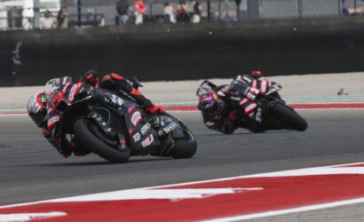 Two motorcycle racers lean into a sharp turn on a racetrack, bikes near ground, close competition with sponsor decals visible on the bikes and suits.