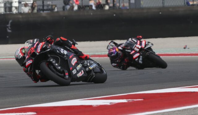 Two motorcycle racers lean into a sharp turn on a racetrack, bikes near ground, close competition with sponsor decals visible on the bikes and suits.