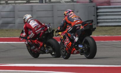 Two motorcycle racers lean into a turn on a racetrack, red and orange livery with sponsor logos visible.