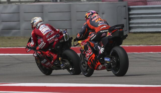 Two motorcycle racers lean into a turn on a racetrack, red and orange livery with sponsor logos visible.