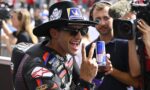Motorsport rider wearing leather suit and hat, sunglasses, holding a Red Bull can while flashing a peace sign. A cameraman films in the background.