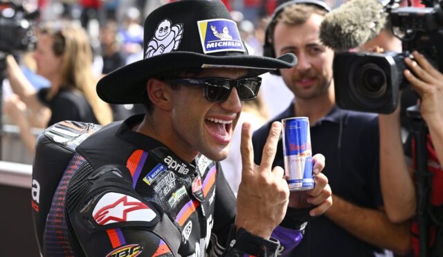 Motorsport rider wearing leather suit and hat, sunglasses, holding a Red Bull can while flashing a peace sign. A cameraman films in the background.
