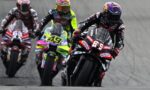 Four motorcycle racers lean into a left-hand turn on a track, with the lead rider wearing #89 in black and red gear and a pink helmet, followed by neon-yellow and red bikes behind.