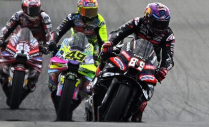 Four motorcycle racers lean into a left-hand turn on a track, with the lead rider wearing #89 in black and red gear and a pink helmet, followed by neon-yellow and red bikes behind.