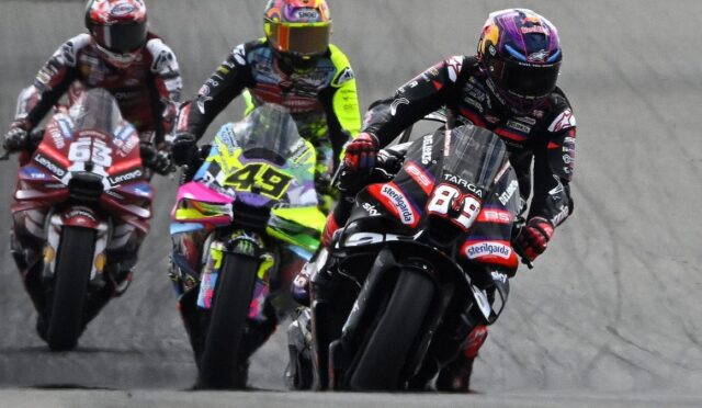 Four motorcycle racers lean into a left-hand turn on a track, with the lead rider wearing #89 in black and red gear and a pink helmet, followed by neon-yellow and red bikes behind.