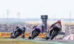 Three motorcycle racers lean into a curve on a race track as they speed through a turn, with a cheering crowd and sponsor banners in the background.