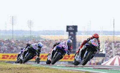 Three motorcycle racers lean into a curve on a race track as they speed through a turn, with a cheering crowd and sponsor banners in the background.