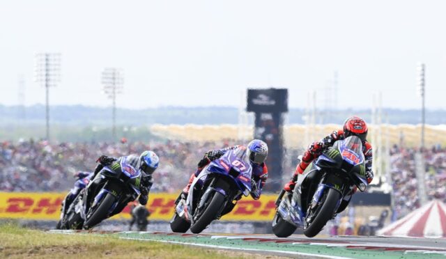 Three motorcycle racers lean into a curve on a race track as they speed through a turn, with a cheering crowd and sponsor banners in the background.