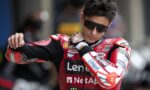 MotoGP rider in a red Lenovo racing suit adjusts his collar while wearing dark sunglasses and a protective glove on one hand.