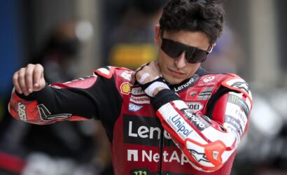 MotoGP rider in a red Lenovo racing suit adjusts his collar while wearing dark sunglasses and a protective glove on one hand.
