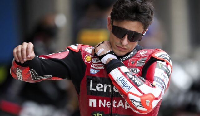 MotoGP rider in a red Lenovo racing suit adjusts his collar while wearing dark sunglasses and a protective glove on one hand.