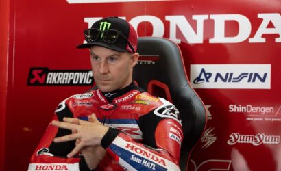 Moto racer in red Honda suit sits in a racing chair with sponsor banners in the background, adjusting his hands.