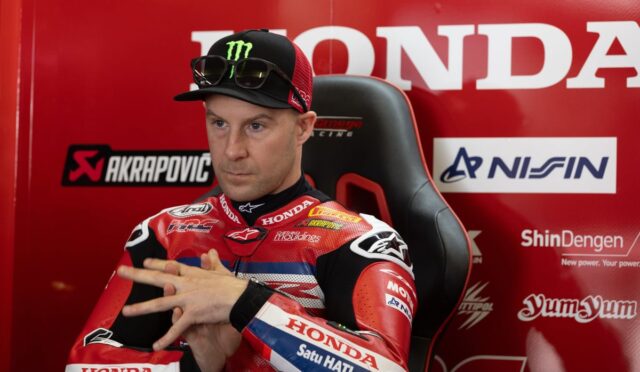 Moto racer in red Honda suit sits in a racing chair with sponsor banners in the background, adjusting his hands.