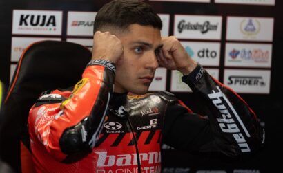 Motorcycle racer in a red racing suit sits in a chair, hands on his head during a press event backdrop of sponsor logos.