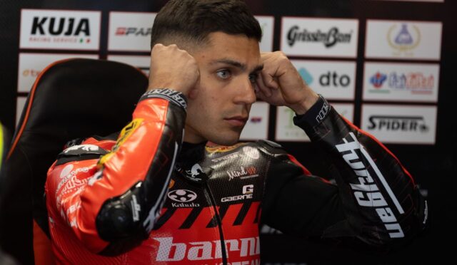 Motorcycle racer in a red racing suit sits in a chair, hands on his head during a press event backdrop of sponsor logos.