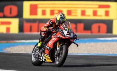 Racing motorcyclist in red gear leaning into a turn on a red Ducati race bike with Pirelli boards in the background.