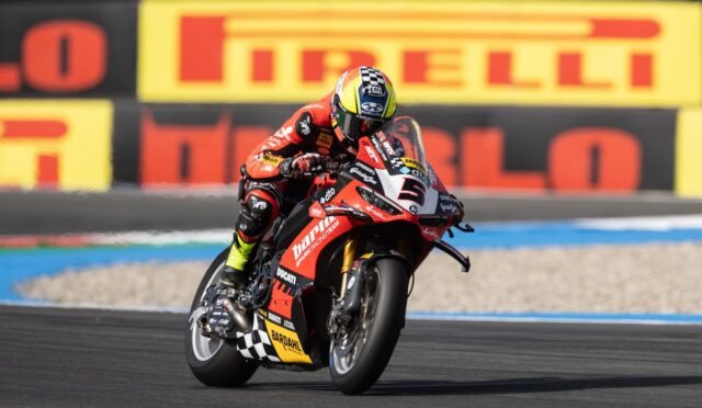 Racing motorcyclist in red gear leaning into a turn on a red Ducati race bike with Pirelli boards in the background.