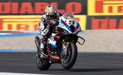 Moto racer performing a wheelie on a racing bike with red, white, and blue livery on a track behind sponsor banners.