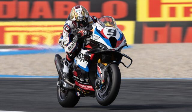 Moto racer performing a wheelie on a racing bike with red, white, and blue livery on a track behind sponsor banners.