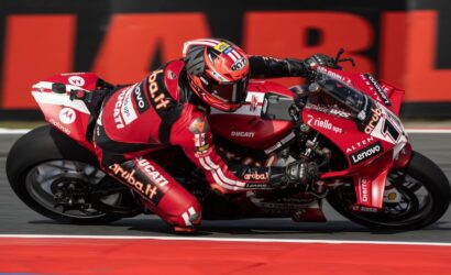 Red Ducati racing bike with sponsor logos leans into a tight turn as a rider in red racing suit hugs the curve on a racetrack with red-white curbs nearby.