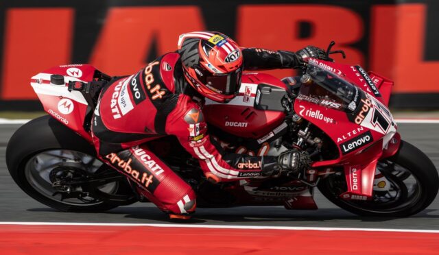 Red Ducati racing bike with sponsor logos leans into a tight turn as a rider in red racing suit hugs the curve on a racetrack with red-white curbs nearby.