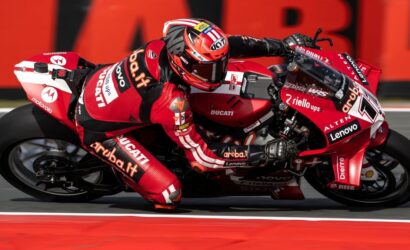 Red Ducati superbike with a rider in a matching red suit leaning hard into a corner on a race track, sponsor logos visible on suit and bike.
