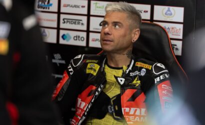 Moto racer with bleached hair sits in a press area, wearing black-red racing leathers with sponsor logos.
