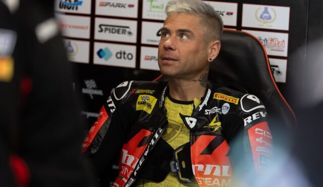 Moto racer with bleached hair sits in a press area, wearing black-red racing leathers with sponsor logos.