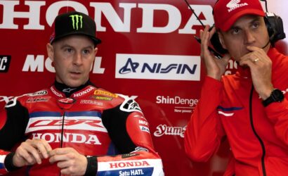 Two motorcycle racers in red Honda racing gear sit in a paddock, one with a Monster cap and the other adjusting a headset.