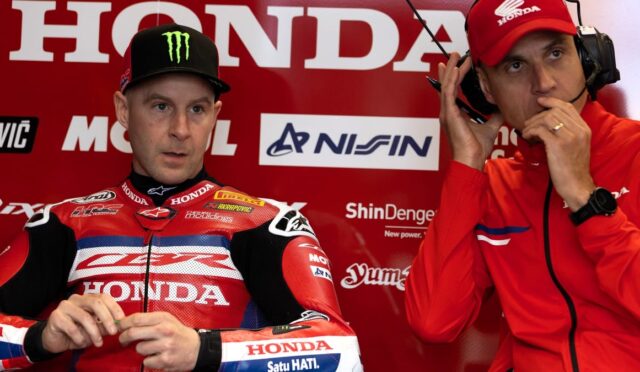 Two motorcycle racers in red Honda racing gear sit in a paddock, one with a Monster cap and the other adjusting a headset.