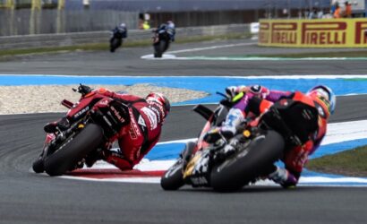 Motorcycle racers lean into a tight turn on a race track, Red bike in foreground and a blue-yellow barrier behind them.