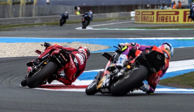 Motorcycle racers lean into a tight turn on a race track, Red bike in foreground and a blue-yellow barrier behind them.