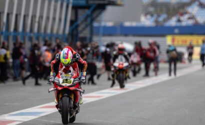 Front-and-center motorcycle racer on a red bike speeds toward the camera, with pit crews and other riders blurred in the background.