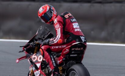 MotoGP rider in a red Ducati racing suit leaning into a turn on a track road surface behind him
