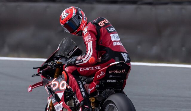 MotoGP rider in a red Ducati racing suit leaning into a turn on a track road surface behind him