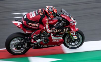 MotoGP rider on a red Ducati racing bike leaning into a corner on a track, wearing a matching red suit with sponsor logos Ariuba.it, Lenovo, and Ducati.