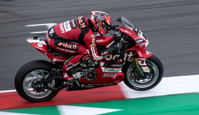 MotoGP rider on a red Ducati racing bike leaning into a corner on a track, wearing a matching red suit with sponsor logos Ariuba.it, Lenovo, and Ducati.
