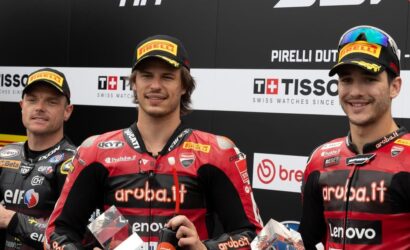 Three motorcycle racers stand on a podium in red-and-black 'aruba.it' racing suits and Pirelli caps, holding trophies against sponsor banners.